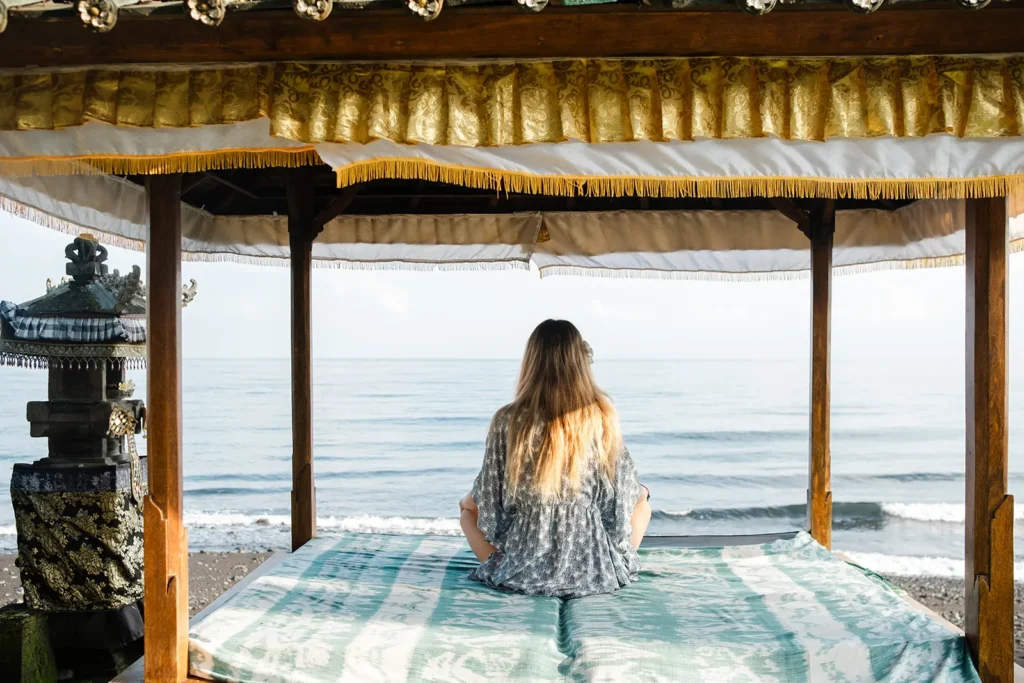 Guest sitting in meditation inside a gold-trimmed Balinese bale facing the sea