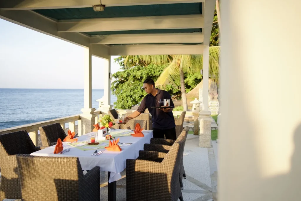 Table set with white linen on the seafront terrace, staff finishing the setting