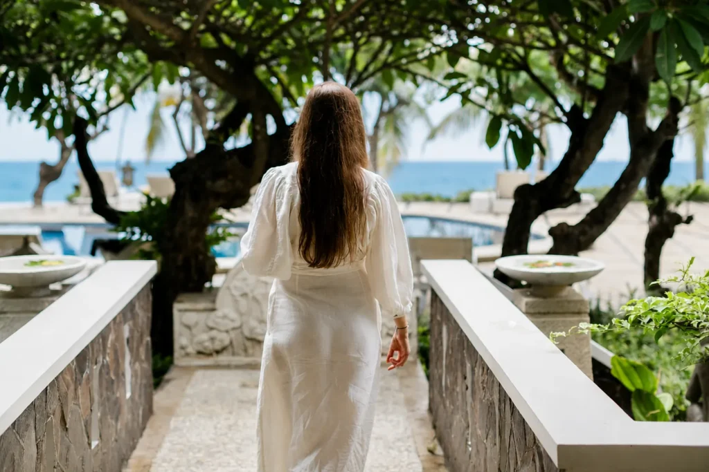 Guest in a white robe walking a garden path toward the pool and the sea